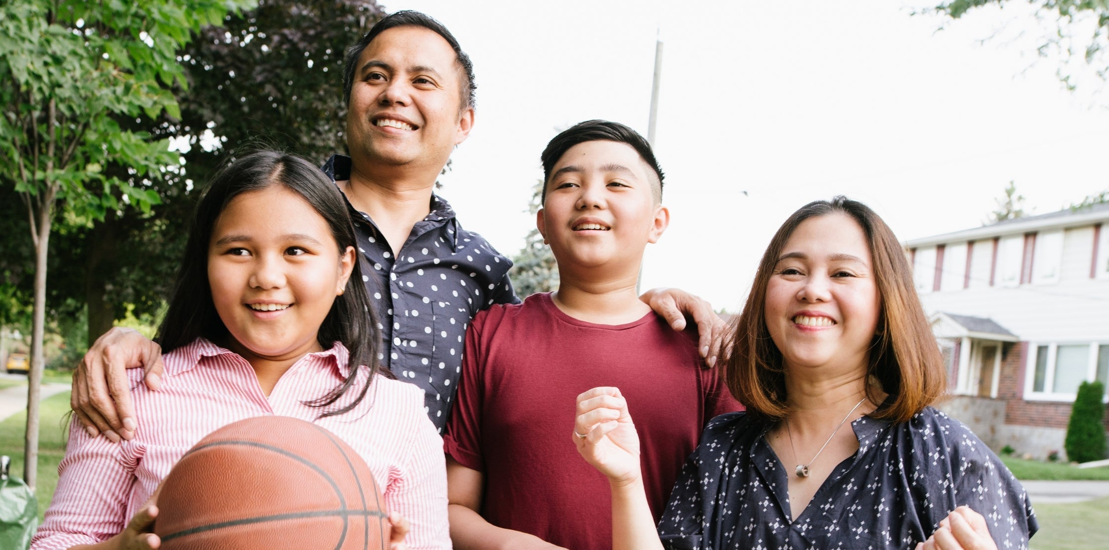 Family of four outdoors with a basketball, smiling at the camera.
