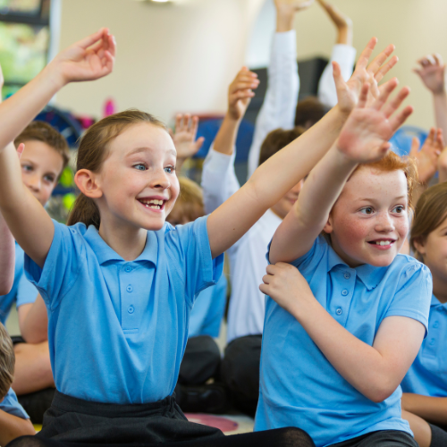 Children in blue polos with raised hands in a classroom setting